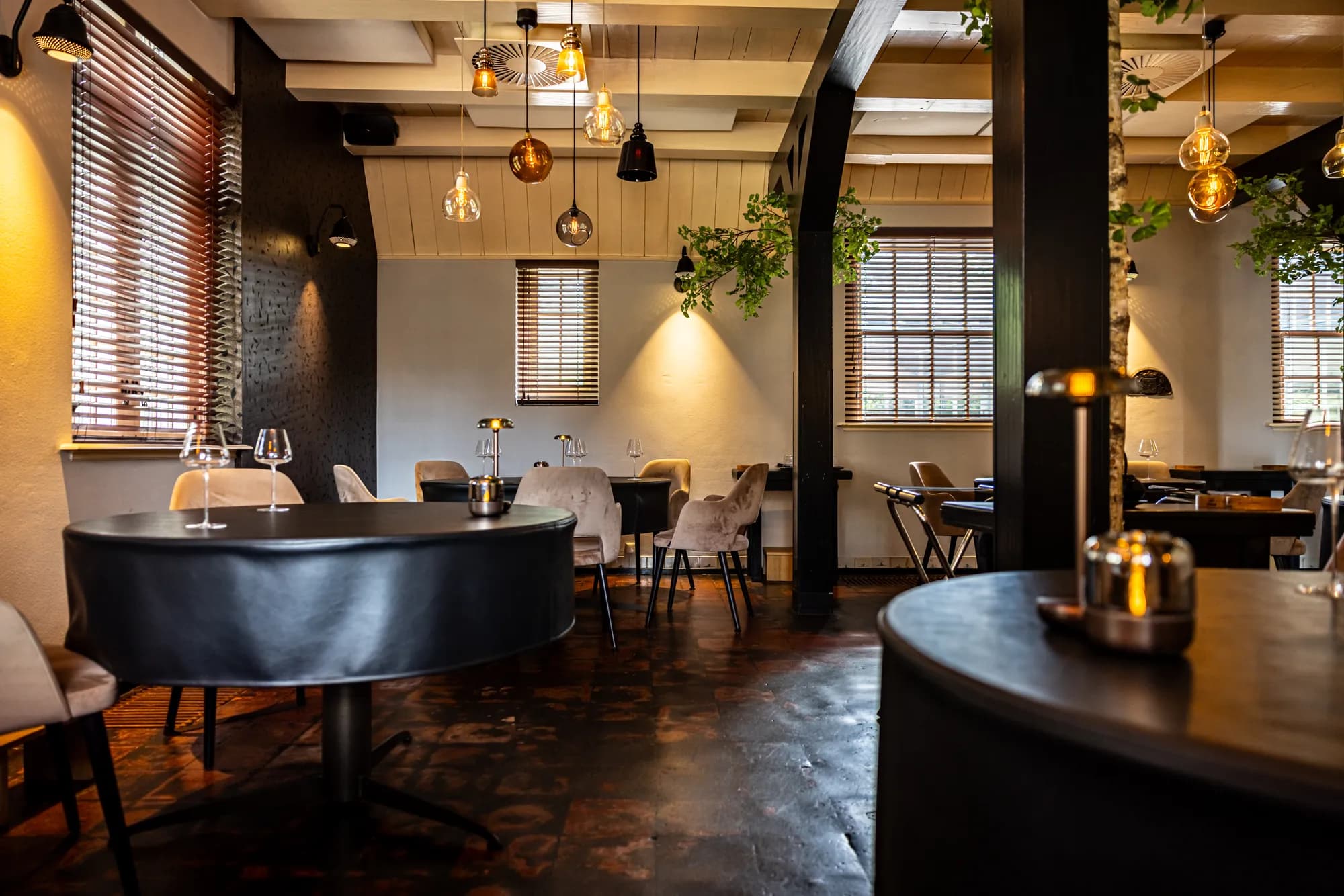De restaurantzaal met ronde tafels, zwarte balken en warme hanglampen
